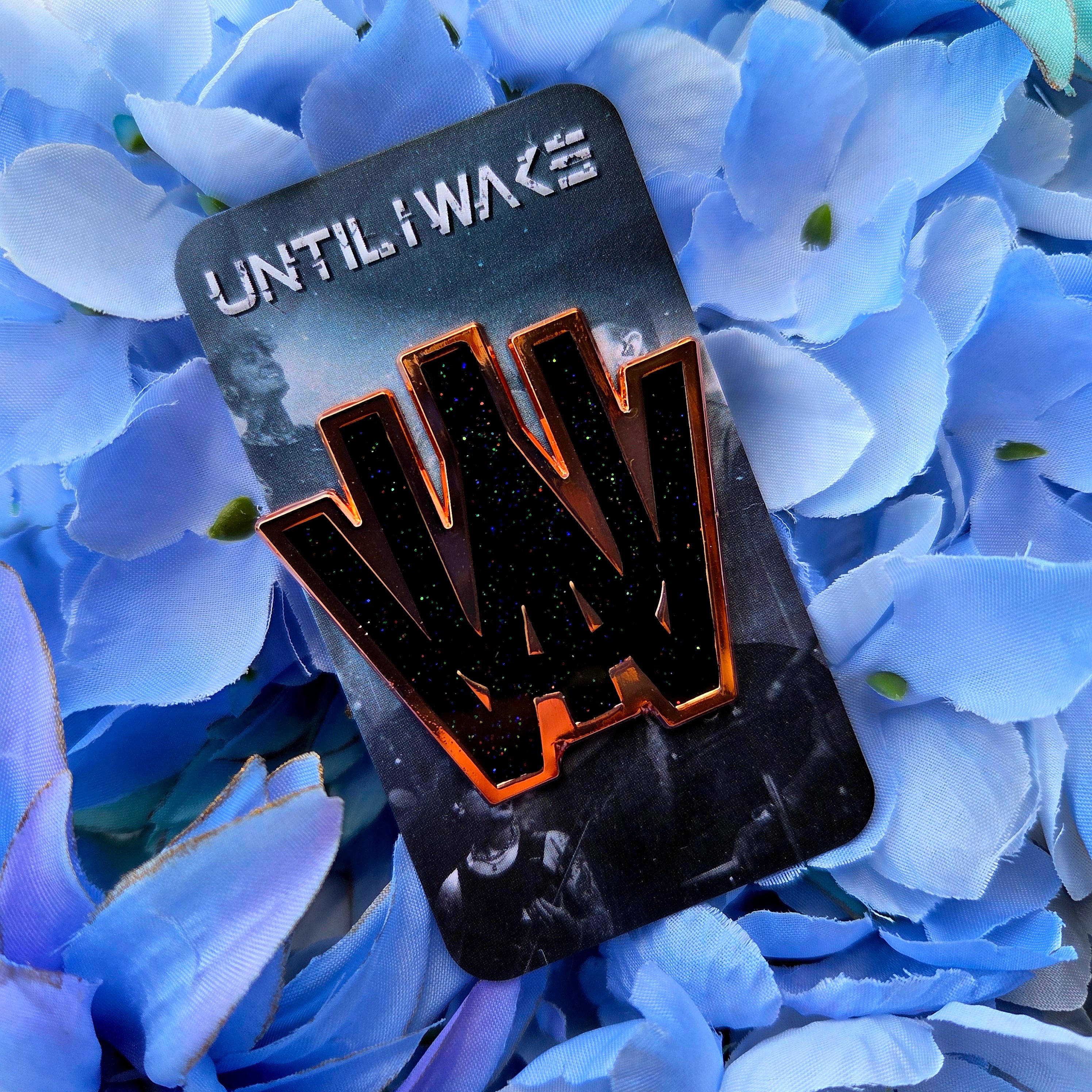 Until I Wake enamel pin on branded backing card showing black glitter pin with copper edges on blue hydrangea petals