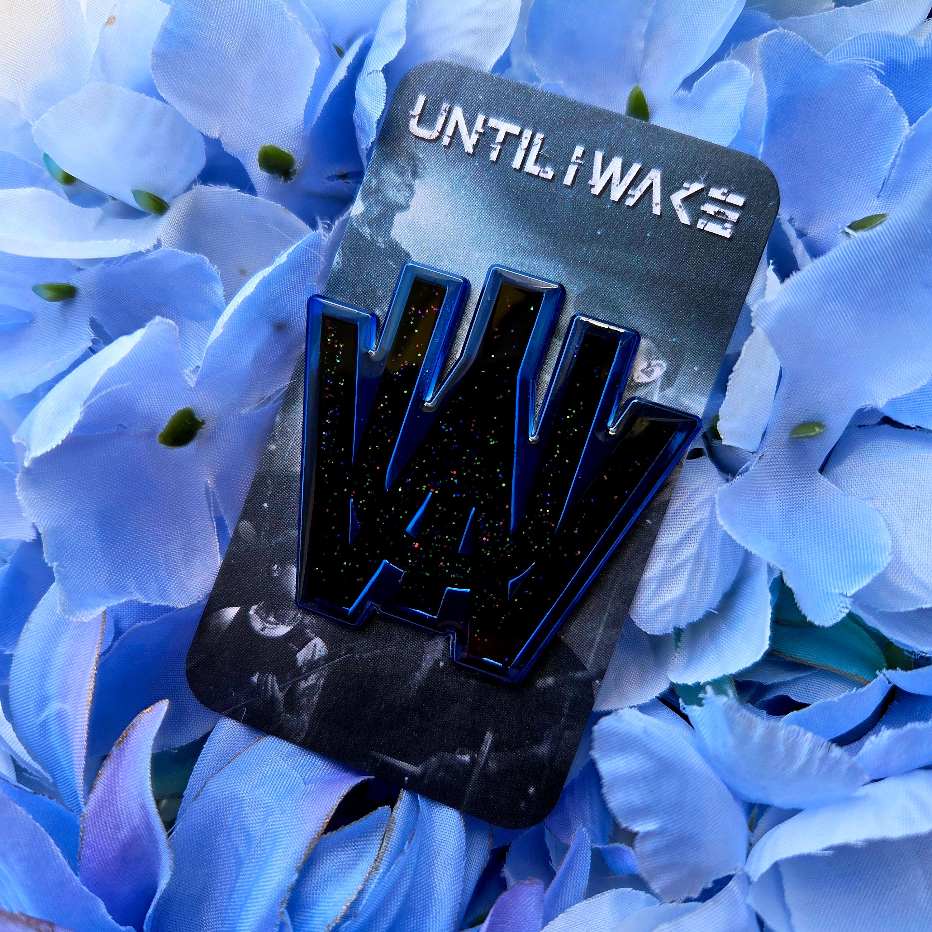 Image of Until I Wake enamel pin by Pincore Merchandise black glitter logo pin on branded backing card against blue flowers