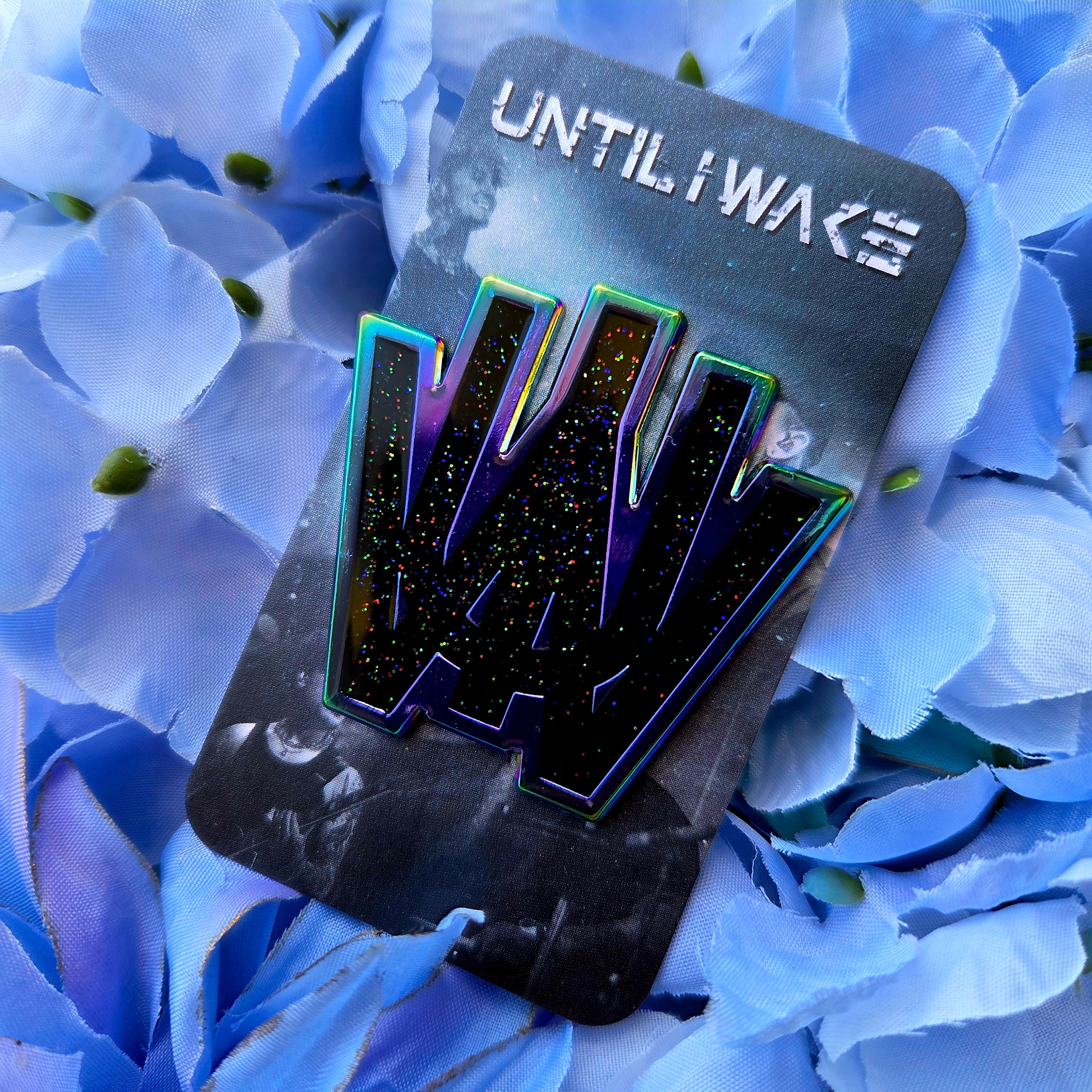Image of Until I Wake enamel pin on Pincore Merchandise card showing black glitter logo with iridescent green edges on blue flower backdrop