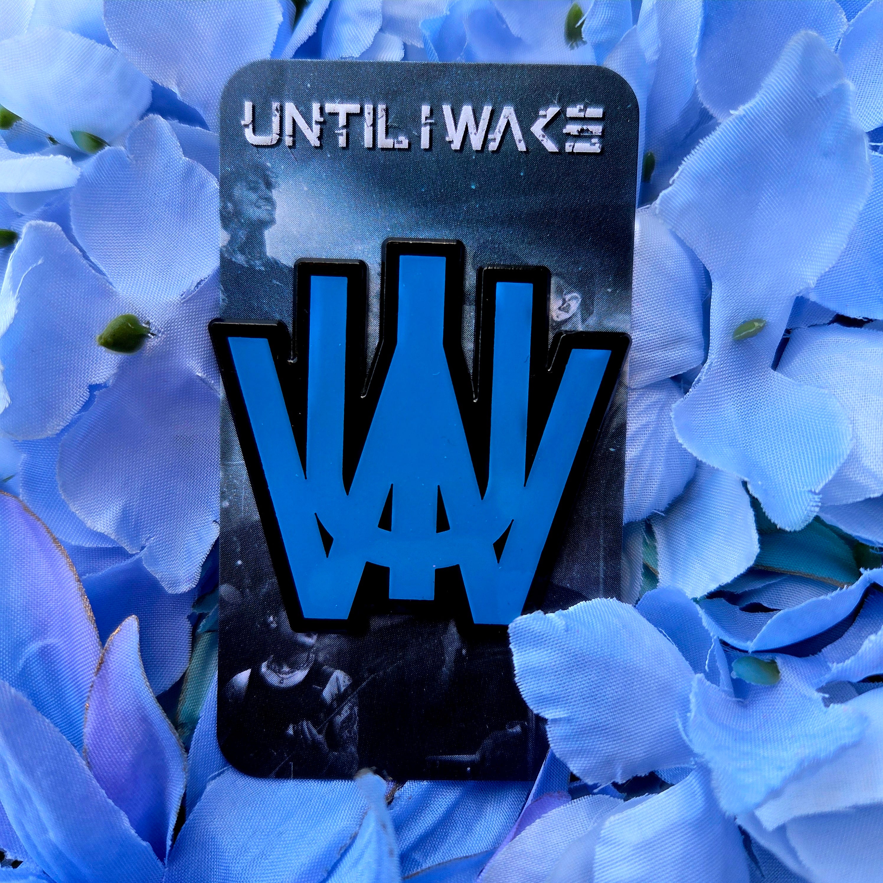 Image of Until I Wake enamel pin featuring blue stylized band logo on black backing card with floral background