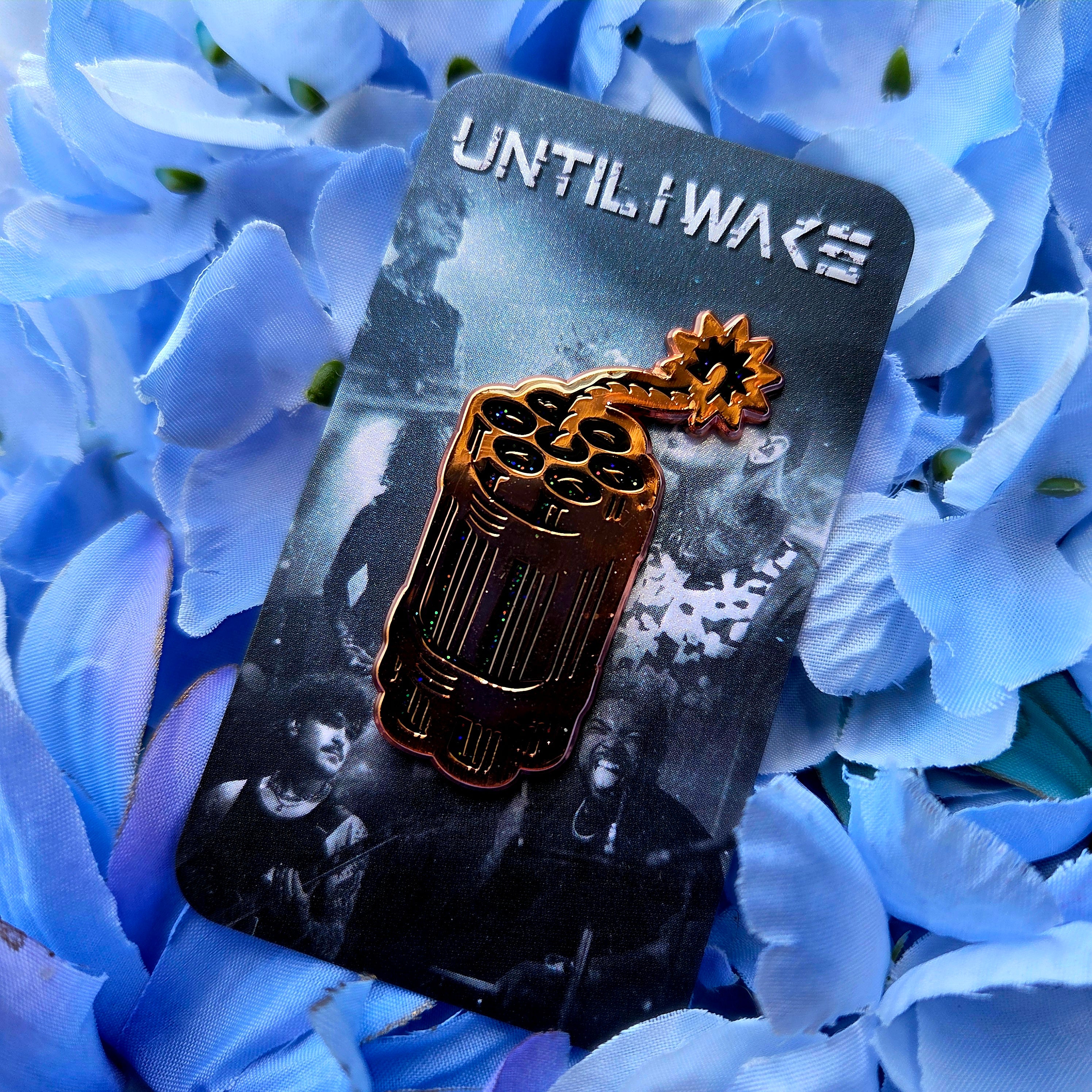 Gold and black Until I Wake enamel pin on black photo backing card featuring band image set on blue flowers